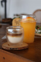 A photo of a coffee and orange juice served in two different glasses on a wooden table. The coffee has a layered appearance with a darker espresso layer on top of a lighter milk layer. The orange juic