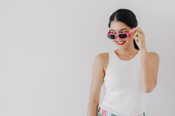 Obraz premium Happy asian Thai woman wear white top and wearing pink sunglasses, chic and fashion, smiling and looking at camera and standing isolated over white background wall.