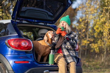 Relaxed woman with dog taking break on road trip, resting with hot tea in car trunk in autumn forest. Female solo traveler enjoys nature views, hiking, exploring camping, outdoor retreat, slow travel