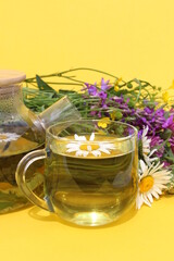 On a yellow background there is a glass cup with chamomile infusion against the backdrop of a bouquet of wild flowers.
