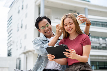 A caring Asian man is placing headphones on his girlfriend while she looks at a tablet.