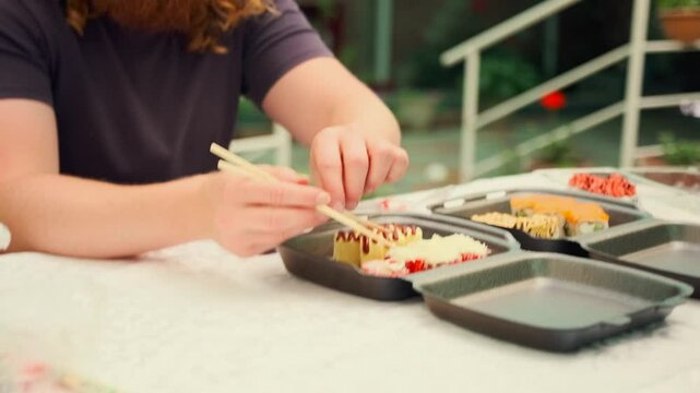 Man hands unsuccessfully tries to take sushi rolls with chopsticks while sitting at a table on the veranda in front of plastic boxes with delivery from a Japanese restaurant.