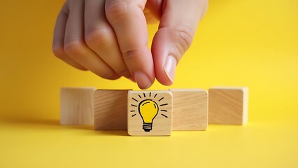 Obraz premium A hand aligns two wooden blocks with a lightbulb drawing on them against a vibrant yellow background. This image symbolizes creativity, innovation