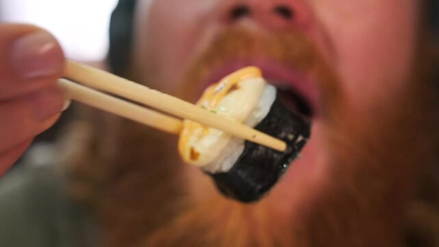 Close up of bearded man putting sushi rolls in mouth using chopsticks with pleasure closing eyes listening to music in big headphones break time.