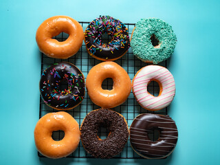 A grid of assorted donuts is displayed on a black cooling rack against a bright turquoise background. The donuts include glazed, chocolate, sprinkle-topped, and coconut-covered varieties.