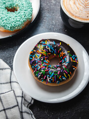 An assortment of donuts is arranged on two plates on a dark, textured surface. A chocolate donut with colorful sprinkles is on a white plate, while another plate has donuts with green sprinkles