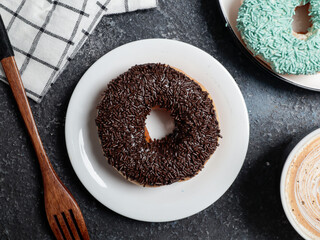 A chocolate-sprinkled donut is placed on a white plate on a dark textured surface. Beside it, a wooden fork rests near a checkered cloth, with a coffee cup and another donut partially visible
