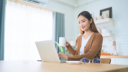 Fototapeta premium Beautiful young Asian woman working with laptop computer in home kitchen. Work at home