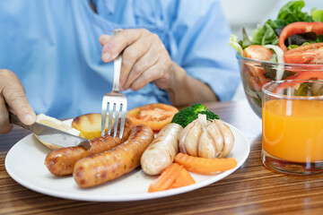 Asian senio woman patient eating breakfast with vegetable healthy food while sitting and hungry on bed in hospital.