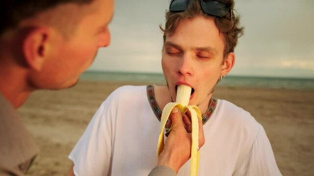 Slow motion, two cute guys eating banana romantically looking into each other's eyes on the beach. Close up.