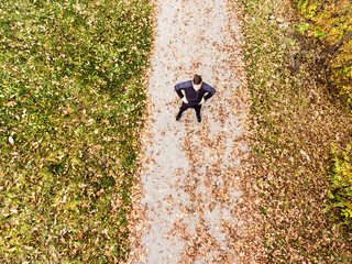 Obraz premium Aerial view of a runner standing on jogging path with fallen leaves and resting after in autumn park. Morning running training.