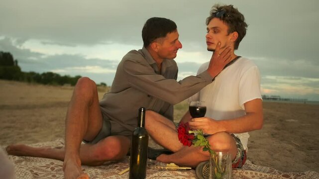 Romantic caresses of two homosexual men on a date with wine and at sunset a couple sitting on the sand near the sea. Slow motion.