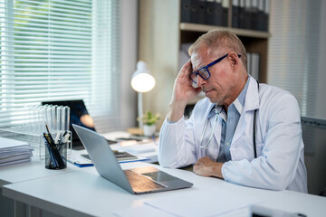 Senior doctor experiencing burnout sitting at his desk looking worried at laptop screen
