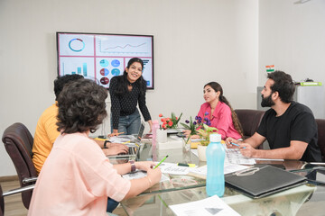 Cheerful Young indian woman gives report or presentation to her colleagues in the Conference Room. showing Graphics, Pie Charts and Company's Growth on the wall tv.