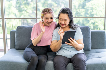 Obraz premium Happy senior asian mother and young daughter waving hands looking at web camera using tablet for video call, smiling mom and girl having fun greeting online by computer webcam making videocall