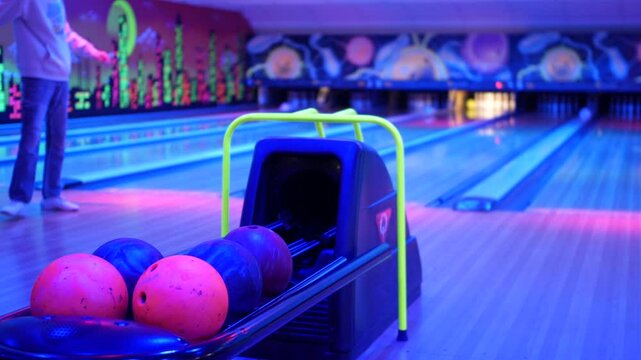 A neon-lit bowling alley with colorful bowling balls lined up in the foreground and a bowler bowling in the background