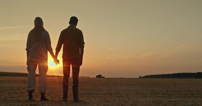 A farmer couple holds hands while admiring the sunset over a field where a combine harvester works in the distance, highlighting the family farm concept.