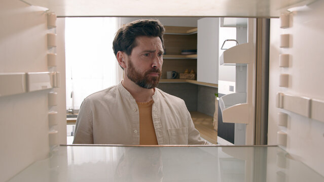 Hungry Caucasian bachelor man looking for food in empty fridge finding no meal products feeling sad disappointed poor unemployment need delivery service POV point of view from inside refrigerator
