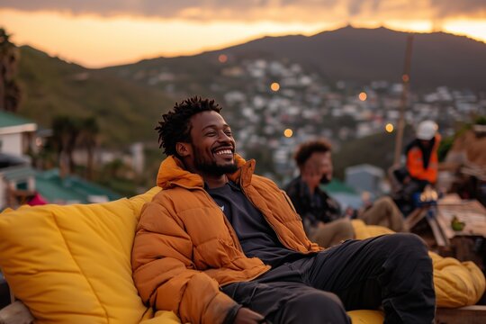 A cheerful man in a warm orange jacket lounges on a comfortable yellow cushion, smiling as he enjoys a peaceful sunset with friends.
