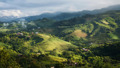 A serene mountain landscape in the Dominican Republic, showcasing lush green hills and valleys. Mist gently rolls over the verdant terrain, with traditional wooden houses dotted throughout.