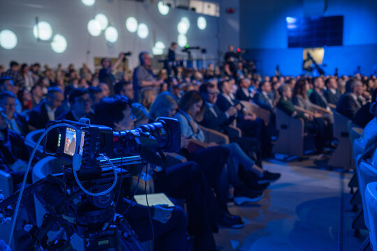 Camera operator capturing live footage of an audience attending a business conference. The room is dimly lit with people seated and focused on the presentation.