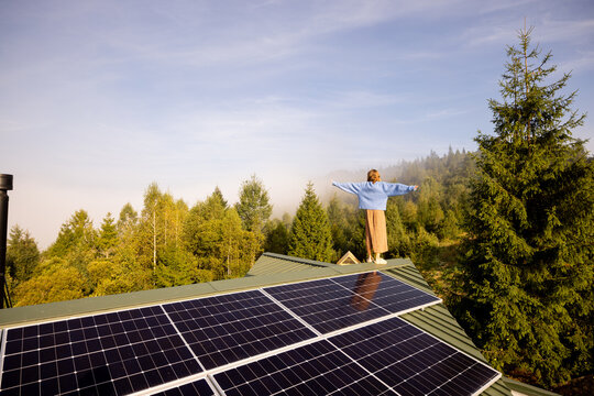 A woman stands on a solar-powered rooftop, arms outstretched, embracing the freedom and sustainability of off-grid living amidst a serene forested landscape
