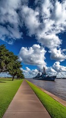 A large gray battleship rests at the pier in Houston, surrounded by greenery, industrial buildings, and a backdrop of blue skies dotted with clouds