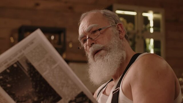 Close-up of bearded senior Caucasian man sitting at table reading newspaper while drinking morning coffee during Christmas holidays season