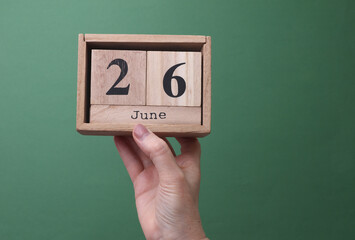 Hand holding a wooden block calendar with the date June 26 on green background