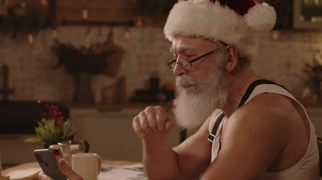 Medium close-up of elderly Caucasian man sitting at table scrolling social networks or reading news in smartphone while having breakfast