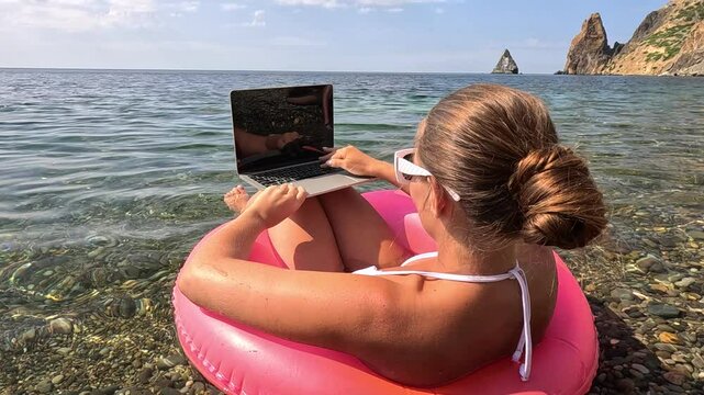 Woman freelancer works on laptop swimming in sea on pink inflatable ring. Happy tourist in sunglasses floating on inflatable donut and working on laptop computer in calm ocean. Remote working anywhere