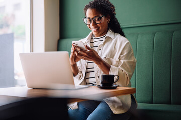 Happy black woman using smartphone with laptop at cafe