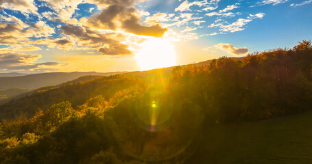 AERIAL, LENS FLARE: Setting sun casting a warm glow over lush autumn forest. Golden light shines across hilly landscape, where vast forests slowly change their shades of green to vibrant fall colors.