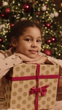 Vertical portrait of pretty young biracial girl posing for camera with big git box. Christmas tree decorated with red and white baubles in background