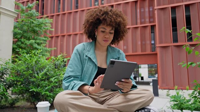 Camera focus on African-American girl sitting on bench in middle of small green zone for relaxation. Using tablet device. Scrolling through everything on screen. Searching for product or information.