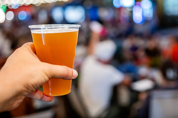 A close-up of a hand holding a plastic cup filled with amber-colored beer, with an out-of-focus background of people in a social setting. The image captures a casual, relaxed atmosphere.