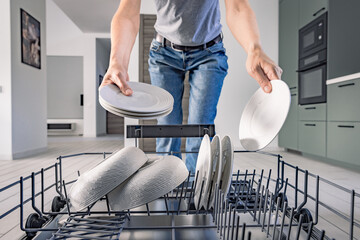 Dishwasher. Young man taking white clear plates from dishwasher. View from dishwasher. Modern kitchen with dishwasher. High quality photo.