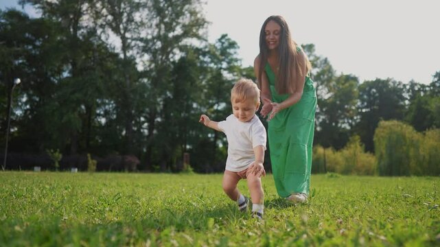 Mom and baby taking the first steps. Happy family kid dream concept. A baby is walking on the grass in a park. Mom and baby walking on lifestyle grass on first steps in the park.