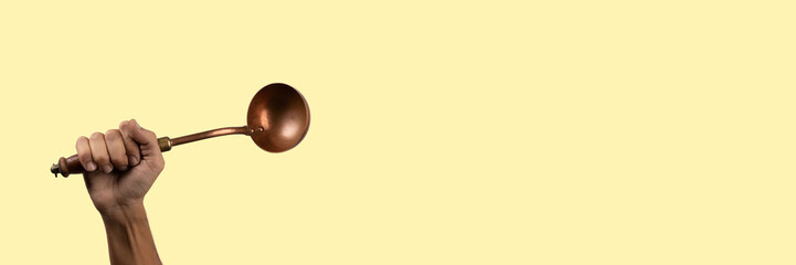 Black male hand holding a brass vintage kitchen ladle on light yellow banner 