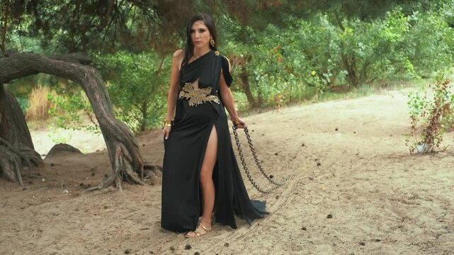 fantasy goth woman holding pulling iron chains stretches across ground. Sexy sorceress Girl witch princess walks through dark forest. long black dress silk train. Concept slavery captive prisoner 4k