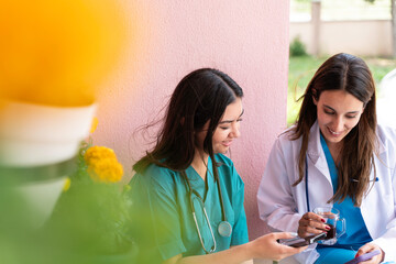 Nurse and female doctor, one in a green scrub top and the other in a white coat, are sitting outside and using cell phones. One woman is holding a cup of coffee.