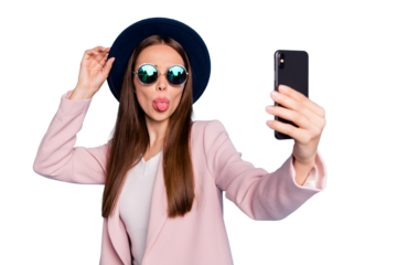 Portrait of playful carefree girl using her cell phone making self photo blogging showing her tongue fooling wearing pink topcoat isolated over blue background