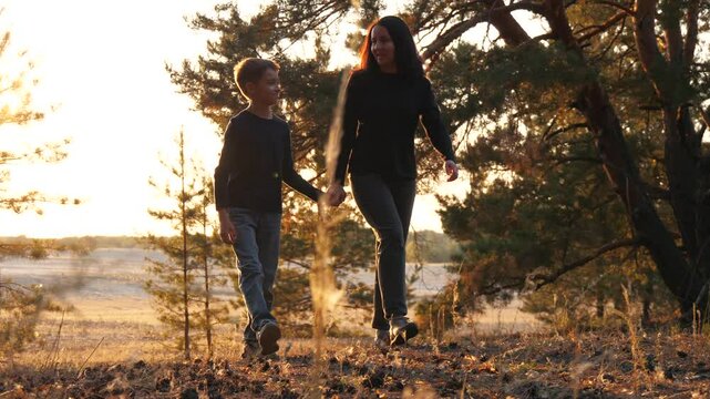 mom and son go autumn forest holding hands. happy boy and mom walk woodland golden hour. fall family vibes in sunset