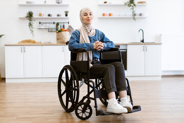 Muslim woman wearing hijab sitting in wheelchair at home in kitchen with modern decor. Woman in...