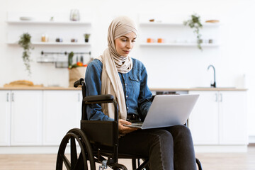 Muslim woman in wheelchair working on laptop in modern kitchen. Confident and focused on her task. Concept of inclusivity, technology, and independence.