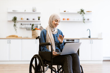 Muslim woman in wheelchair using laptop in home kitchen. Bright, modern kitchen setting with woman smiling and interacting on laptop. Concept of technology, accessibility, independence, and home.