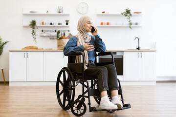 Muslim woman in wheelchair talking on phone in modern home kitchen. Shows independence, daily life, positivity. Background features clean, minimalist kitchen with white cabinets, green plants.