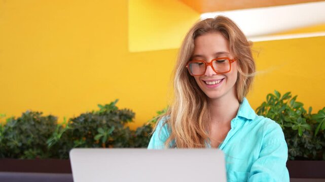 Happy female developer in trendy eyeglasses working on laptop at cafe. Woman freelancer is coding at restaurant on laptop. Caucasian girl is working remotely. 