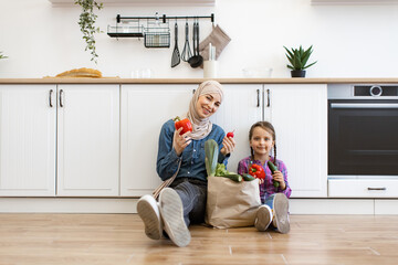 Muslim mother and daughter sit in modern kitchen preparing to cook healthy food together. They are surrounded by fresh vegetables promoting family bonding and healthy lifestyle.