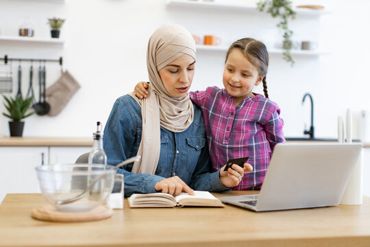 Captures family bond, online shopping convenience and cultural representation. Muslim mother and daughter sitting at kitchen table and buying ingredients for meal using laptop, bank card, recipe book